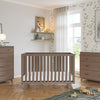 A modern nursery features a gray crib, dresser with the Child Craft F01792 Universal Changing Table Topper in Dusty Heather, wooden toys on a green rug, light wood floors, green leaf wallpaper, decor shelves, and a large window.