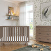 A modern nursery with a wooden crib, dresser fitted with the Child Craft F01792 Universal Changing Table Topper in Dusty Heather, toy blocks and trucks on a green rug, leafy wallpaper, shelves with books and decor, potted plant, and sunlight through sheer curtains.