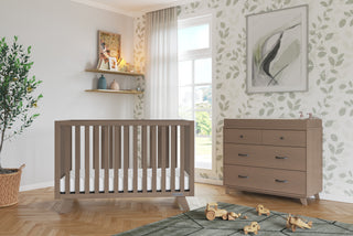A modern nursery with a wooden crib, dresser fitted with the Child Craft F01792 Universal Changing Table Topper in Dusty Heather, toy blocks and trucks on a green rug, leafy wallpaper, shelves with books and decor, potted plant, and sunlight through sheer curtains.