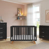 A modern nursery featuring a black crib, a matching dresser topped with the Child Craft F01792 Universal Changing Table Topper in Ebony, soft pink walls, sheer curtains, wood accents, and a decor shelf above the crib.