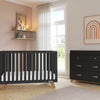 A modern nursery features a black crib, matching dresser with the Child Craft F01792 Universal Changing Table Topper in Ebony, wooden floors, light pink walls with shelves, framed rainbow art, and a large window with sheer curtains letting in natural light.