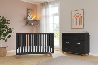 A modern nursery features a black crib, matching dresser with the Child Craft F01792 Universal Changing Table Topper in Ebony, wooden floors, light pink walls with shelves, framed rainbow art, and a large window with sheer curtains letting in natural light.