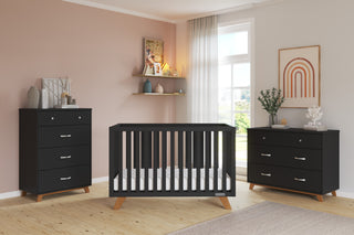 Modern nursery with a pink accent wall, Child Craft Soho 4-in-1 Convertible Crib in Ebony at the center, matching dressers on each side, large sunlit window, plus shelves, plants, and vibrant wall art for décor.