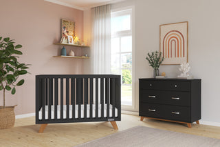 A modern nursery with a black crib, wooden floor, blush pink accent wall, shelves with decor, a window with sheer curtains, and framed rainbow art above the Child Craft Soho 3-Drawer Dresser in Ebony.