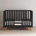 The Child Craft Soho 4-in-1 Convertible Crib in Ebony features vertical slats, wooden legs, a white mattress, and partial side rails. Its mid-century modern design stands out on a light wood floor against a plain white wall.