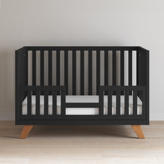 The Child Craft Soho 4-in-1 Convertible Crib in Ebony features vertical slats, wooden legs, a white mattress, and partial side rails. Its mid-century modern design stands out on a light wood floor against a plain white wall.