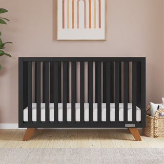 The Child Craft Soho 4-in-1 Convertible Crib in Ebony, featuring wooden legs, stands against a pink wall with abstract art above. A plant is on the left and woven baskets with blankets are on the right, atop a light wood floor with a rug.