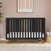 The Child Craft Soho 4-in-1 Convertible Crib in Ebony, featuring wooden legs, stands against a pink wall with abstract art above. A plant is on the left and woven baskets with blankets are on the right, atop a light wood floor with a rug.