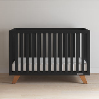 The Child Craft Soho 4-in-1 Convertible Crib in Ebony showcases a modern black finish, wooden legs, vertical slats, and a white mattress—set on a light wood floor against a plain white wall.