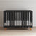 The Child Craft Soho 4-in-1 Convertible Crib in Ebony showcases a modern black finish, wooden legs, vertical slats, and a white mattress—set on a light wood floor against a plain white wall.