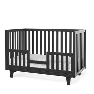 The Child Craft F09544 Toddler Guard Rail in Ebony features open slats on all sides, a low entry guard rail for safety, and a white patterned mattress inside. Crafted as a non-toxic baby crib, it combines style with peace of mind.