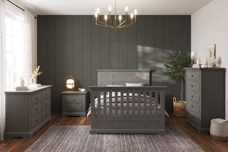 A modern bedroom with dark paneled walls, crown molding, a gray wood bed, dresser and chest, Child Craft Jordyn Nightstand in Lunar Gray (ready-to-assemble), gray rug, wood floor, chandelier, plants and books for decor. Natural light enters from a window.