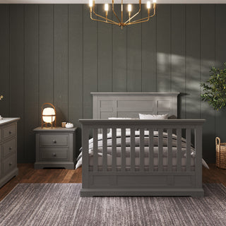 A modern bedroom features a gray crib-style bed, baby-safe finishes, a Child Craft Jordyn Nightstand in Lunar Gray with a lamp, gray rug on wooden floors, dark paneled walls with crown molding, a chandelier, and a potted plant in the corner.