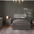 A modern bedroom features a gray crib-style bed, baby-safe finishes, a Child Craft Jordyn Nightstand in Lunar Gray with a lamp, gray rug on wooden floors, dark paneled walls with crown molding, a chandelier, and a potted plant in the corner.