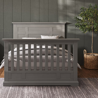 A Child Craft Jordyn Nightstand in Lunar Gray stands ready-to-assemble next to a bed against a dark paneled wall with crown molding, with a potted plant in a wicker basket nearby on a patterned rug.