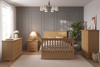 A modern bedroom with light wood furniture, including a bed, dresser, chest, and the Child Craft Jordyn Nightstand (Ready-to-Assemble, Biscotti). A gray paneled wall, large window, plant, lamp, and minimalist decor complete the space.