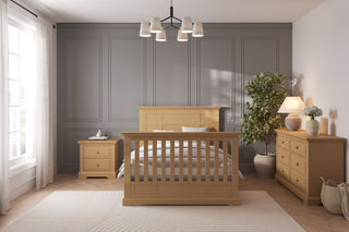 A modern bedroom features a gray paneled wall, wooden bed frame, and a Child Craft Jordyn Nightstand in Biscotti. A large plant, table lamp, dresser, and sheer white curtains on the window let in plenty of natural light.