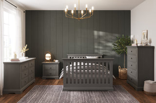 A modern bedroom with dark wood furniture—bed, dresser, chest, and the Child Craft Jordyn Nightstand in Lunar Gray. Features include a dark accent wall, gold chandelier, crown molding, large window with white curtains, and a gray area rug.