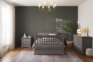 A modern bedroom with crown molding, a dark paneled accent wall, gray wood bed, Child Craft Jordyn Nightstand in Lunar Gray, dresser, large window with white curtains, potted plant, gold chandelier, and a gray rug on the wooden floor.