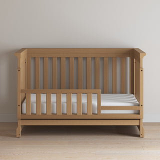 The Child Craft F09554 Toddler Guard Rail in Biscotti features vertical slats and a partial crib guard rail, shown against a plain light-colored wall on a light wood floor.