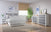 A bright, modern bedroom with white and gray furniture, featuring Child Craft Full-Size Bed Rails in white/brown, a nightstand, dressers, a large green plant by the window, abstract wall art, and a beige rug on a wooden floor.