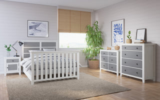 A modern bedroom features Child Craft’s Ocean Grove Nightstand in white/gray, with matching furniture, wood flooring, a large rug, wall art, a potted plant, and a window with bamboo blinds that let in natural light.