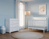 A serene nursery with pale blue walls, the Child Craft Scout 4-in-1 Convertible Crib in matte white, a white changing table with drawers, a diaper bin, beige floor lamp, sheer curtains on a tall window, and framed leaf art above the crib.