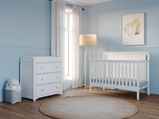 A serene nursery with pale blue walls, the Child Craft Scout 4-in-1 Convertible Crib in matte white, a white changing table with drawers, a diaper bin, beige floor lamp, sheer curtains on a tall window, and framed leaf art above the crib.