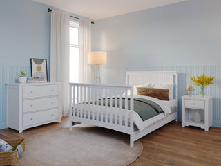 A cozy bedroom features the Child Craft Harmony 3-Drawer Dresser with Changing Table Topper in matte white, matching bed and nightstand, a lamp, sheer curtains, rug, and flowers. Light blue walls and wood floors make it perfect for nursery storage and comfort.