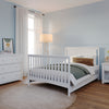 A cozy bedroom features a Child Craft Scout 4-in-1 Convertible Crib in Matte White, a matching white bed, nightstand with yellow flowers, dresser, floor lamp, sheer-curtained window, round beige rug, all set against light blue walls.