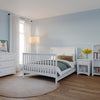 A cozy bedroom with light blue walls, white baby nursery furniture including the Child Craft Scout 4-in-1 Convertible Crib in Matte White, dresser, nightstand, bookshelf, a round rug, window with sheer curtains, floor lamp, and decorative accents.