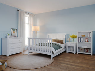 A cozy bedroom with light blue walls, white baby nursery furniture including the Child Craft Scout 4-in-1 Convertible Crib in Matte White, dresser, nightstand, bookshelf, a round rug, window with sheer curtains, floor lamp, and decorative accents.