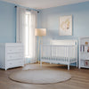 A bright, modern nursery with light blue walls, the Child Craft Scout 4-in-1 Convertible Crib in matte white, matching dresser and bookshelf, a beige rug, floor lamp, window with white curtains, and a framed animal print on the wall.