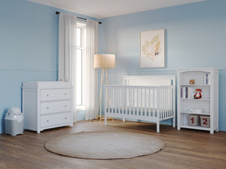 A bright, modern nursery with light blue walls, the Child Craft Scout 4-in-1 Convertible Crib in matte white, matching dresser and bookshelf, a beige rug, floor lamp, window with white curtains, and a framed animal print on the wall.