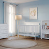 A bright nursery with light blue walls features the Child Craft Scout 4-in-1 Convertible Crib in matte white, a dresser, bookshelf, floor lamp, white curtains, and a round beige rug on a wooden floor, with framed botanical art above the crib.