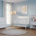 A bright nursery with light blue walls features the Child Craft Scout 4-in-1 Convertible Crib in matte white, a dresser, bookshelf, floor lamp, white curtains, and a round beige rug on a wooden floor, with framed botanical art above the crib.
