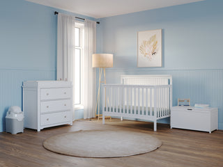 A bright nursery with light blue walls, a white crib and dresser, the Child Craft Harmony 33" Kids Toy Box in matte white, a large window with white curtains, floor lamp, framed wall art, diaper pail, and round beige rug on wooden floors.