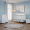 A serene baby nursery with light blue walls, white furniture including the Child Craft Scout 4-in-1 Convertible Crib in Matte White, dresser, toy chest, a beige round rug, standing lamp, window with white curtains, and framed feather artwork.