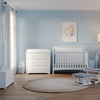 A bright nursery with light blue walls features the Child Craft Scout 4-in-1 Convertible Crib in Matte White, a dresser, rocking chair with ottoman, storage chest, round beige rug, floor lamp, and framed leaf artwork. Sunlight streams through the window.