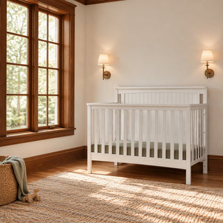 A white Child Craft Scout 4-in-1 Convertible Crib with a solid beadboard headboard sits in a room with a window and two wall sconces.