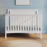 The Child Craft Scout 4-in-1 Convertible Crib in Matte White, featuring vertical slats, stands against a blue paneled wall. A light rug lies on the wooden floor, creating a cozy baby nursery with a framed picture above the crib.