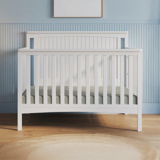 The Child Craft Scout 4-in-1 Convertible Crib in Matte White, featuring vertical slats, stands against a blue paneled wall. A light rug lies on the wooden floor, creating a cozy baby nursery with a framed picture above the crib.