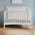 The Child Craft Scout 4-in-1 Convertible Crib in Matte White, featuring vertical slats, stands against a blue paneled wall. A light rug lies on the wooden floor, creating a cozy baby nursery with a framed picture above the crib.