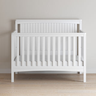 The Child Craft Scout 4-in-1 Convertible Crib in Matte White, featuring vertical slats, stands on a light wood floor against a beige wall, creating a cozy, neatly-made focal point for any baby nursery.