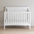 The Child Craft Scout 4-in-1 Convertible Crib in Matte White, featuring vertical slats, stands on a light wood floor against a beige wall, creating a cozy, neatly-made focal point for any baby nursery.