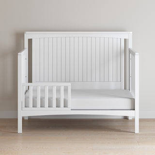 The Child Craft Scout 4-in-1 Convertible Crib in Matte White features a partial safety rail and wooden slat paneling, making it a minimalist nursery piece that pairs well with light walls and wooden floors.
