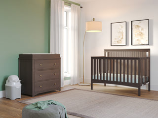 A modern nursery with green and white walls features the Child Craft Scout 4-in-1 Convertible Crib in Dapper Gray, matching changing table, floor lamp, two framed leaf prints, a white diaper bin, and a light rug on wood flooring.