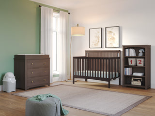 A cozy nursery with a green accent wall, crib, dresser with changing pad, Child Craft Harmony 4-Shelf Bookcase in Dapper Gray, storage bins, lamp, pouf, and large window with white curtains. Light wood floor and a neutral rug complete the room.