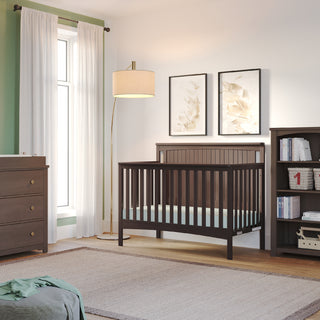 A cozy nursery features a dark brown crib, matching dresser, and the Child Craft Harmony 4-Shelf Bookcase in Dapper Gray. A floor lamp sits by the window with white curtains, two botanical prints hang above the crib, and a beige rug covers the wooden floor.