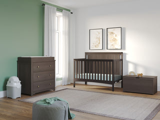 A modern nursery with a green accent wall, dark wood crib, and the Child Craft Harmony 3-Drawer Dresser with Changing Table Topper in Dapper Gray. A diaper pail sits by the dresser, light rug on the floor, two botanical prints above the crib, and natural light fills the room.